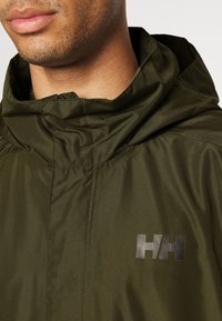 Veste à capuche verte olive en tissu lisse, ornée d'un logo noir sur la poitrine et d'un col haut pour une protection accrue.