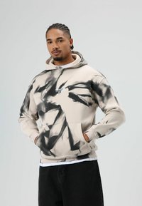 Beige capuchontrui met zwart tie-dye patroon, kangoeroezak en geborduurde tekst. Draagt over een wit T-shirt en zwarte broek.