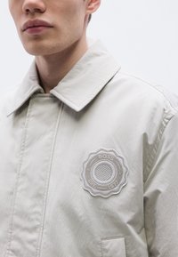 Μπεζ ανοιχτόχρωμο σακάκι με κλείσιμο με κουμπιά μπροστά, φαρδύ γιακά και υφασμάτινο σήμα με ανάγλυφη υφή. Λείο ύφασμα με ορατές λεπτομέρειες ραφής.