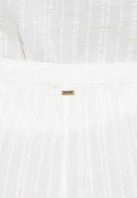 Hvidt stof med et tekstureret stregmønster, udstyret med en elastisk talje og en lille guldfarvet "Rip Curl" mærkat.