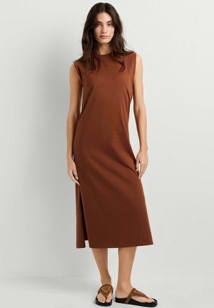 Vrouw die een mouwloze, bruine midi-jurk met zijsplit en bruine platte sandalen draagt, staande tegen een effen lichtgrijze achtergrond.