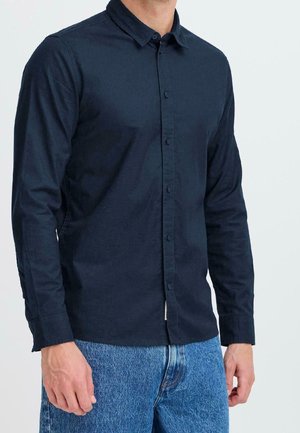 Man draagt een donkerblauw overhemd met knopen en lange mouwen en een blauwe spijkerbroek, staand tegen een effen lichte achtergrond.