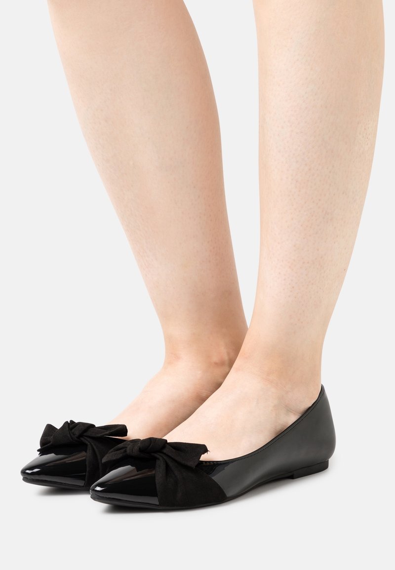 Pieds chaussés de ballerines en cuir verni noir avec de grands nœuds en tissu sur les orteils sur un fond neutre.