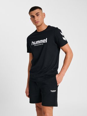 Jonge man die zwarte hummel sportkleding met witte logo's draagt, staand met één hand in de zak tegen een effen lichte achtergrond.