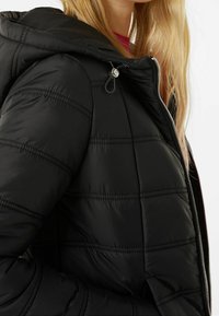 Czarna puchowa kurtka z przeszywanymi panelami, zamkiem błyskawicznym i kapturem ze sznurkiem. Tkanina wydaje się miękka i błyszcząca.