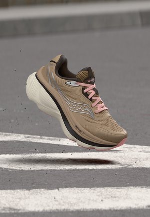 Chaussure de course beige et rose Saucony avec une semelle épaisse en plein air au-dessus d'une surface goudronnée grise avec des marquages blancs.
