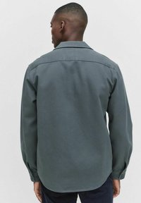 Donkergroen, langemouwen shirt met een dichtgeknoopte kraag en een afgeronde zoom. Gemaakt van een gladde stof, met zichtbare stiksels en knopen bij de manchetten.