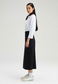 Vit oversized långärmad skjorta, svart midi kjol och vita sneakers med gröna detaljer; svart prickig scarf runt halsen.