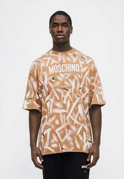 Ενήλικας που φοράει ένα μπεζ, πολύχρωμο t-shirt με μοτίβο πινελιάς Moschino Milano και μαύρο παντελόνι Moschino, όρθιος μπροστά σε ένα απλό φόντο.