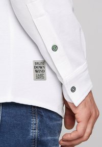 Camicia bianca a maniche lunghe con tessuto texturizzato, dotata di un'etichetta cucita con la scritta "SAILING DOWN WIND CAMP DAVID" e polsini con bottoni.