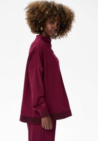 Vrouw met krullend haar, gekleed in een oversized bordeauxrode top en bijpassende broek, staande zijwaarts tegen een witte achtergrond.