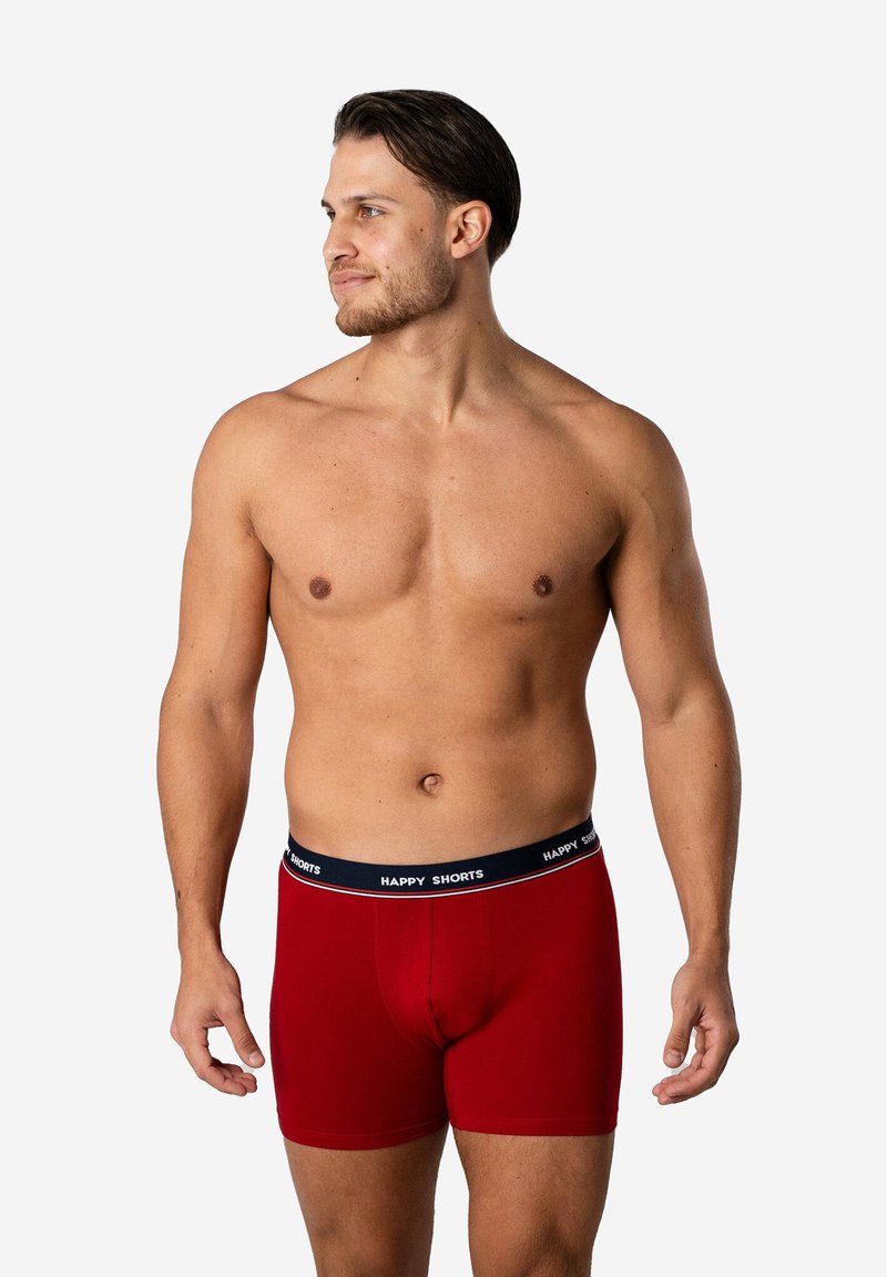 Červené boxerky vyrobené z hladké tkaniny, s černým elastickým pasem na kterém je vytištěno "HAPPY SHORTS" v bílé barvě.