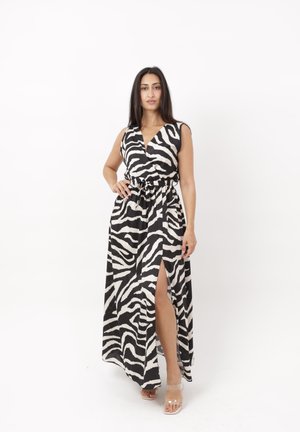 Vrouw in mouwloze zwart-wit zebraprint jurk met split aan de zijkant, staand met één hand op de heup tegen een witte achtergrond.