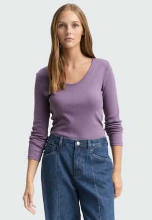 Młoda kobieta z długimi blond włosami, ubrana w dopasowaną fioletową bluzkę z długim rękawem i niebieskie jeansy z wysokim stanem, stojąca na jednolitym tle.