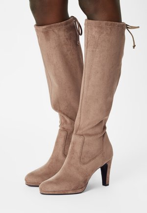 Bottes hautes en daim taupe à talons empilés et bouts arrondis, portées sur des jambes avec des fermetures à cordon coulissant au sommet à l'arrière.