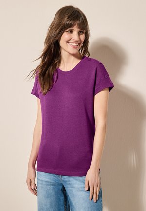 Top corto de color púrpura de manga corta en tejido suave, con un cuello redondo y detalles de botones en los hombros, combinado con unos vaqueros de color azul claro.