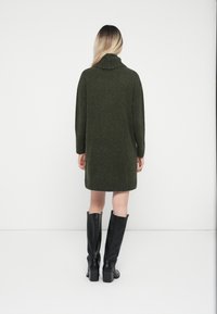 Knälång, mörkgrön tröjdress med hög krage och långa ärmar, matchad med svarta, knähöga klackstövlar. Slät textur.