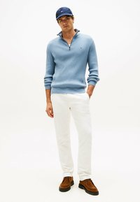 Lichtblauwe ribgebreide trui met een ritssluiting op de kraag, witte broek en bruine schoenen. Een marineblauwe pet maakt de outfit compleet. Eenvoudig, casual ontwerp.