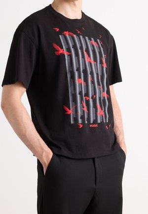 Person iført en sort T-shirt med lodret grå tekst og røde fugle-grafik, hænderne i lommerne, stående mod en ensfarvet lys baggrund.