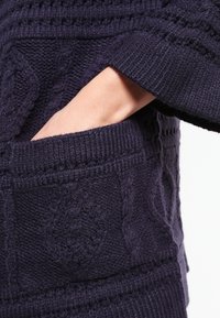 Mörkblå stickad tröja med kabelmönster, som har en ficka och ribbade ärmslut. Texturen verkar mjuk och tjock, vilket erbjuder värme och komfort.