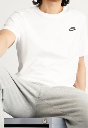 Personne portant un t-shirt blanc Nike et un pantalon de jogging gris, assise sur un casier ou une armoire métallique grise, devant un fond uni.