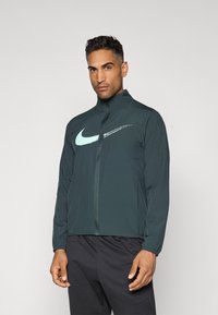Nike-takki tummanvihreänä, jossa on korkea kaulus, vetoketjusulku ja vaaleansininen logo. Made from smooth, lightweight material.