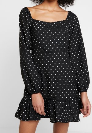 Zwarte jurk met lange opgeblazen mouwen, vierkante halslijn en wit sterrenpatroon. Heeft een gerimpelde zoom en een lichte stoftextuur.