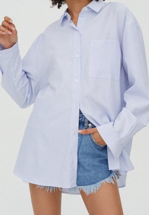 Femme portant une chemise oversize à rayures bleu clair et blanches, boutonnée, par-dessus un short en jean effiloché, main dans la poche, sur fond uni.