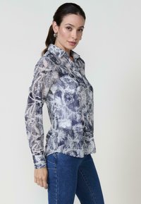 Una donna posa di profilo, indossando una camicia a maniche lunghe con motivo e jeans. I suoi capelli sono raccolti in una coda di cavallo, mettendo in mostra orecchini a cerchio.