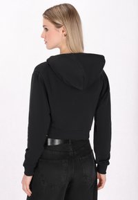 Sudadera negra recortada con capucha, mangas largas y puños acanalados, combinada con pantalones negros de talle alto con bolsillos traseros.