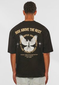 Sort oversized t-shirt med et hvidt duegrafik, teksten "Rise Above the Rest" og guld-accenter. Korte ærmer og en afslappet pasform.