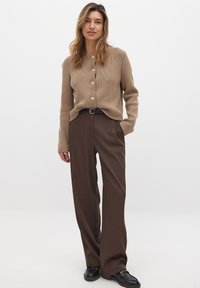 Cardigan en tricot beige avec un motif texturé et un col rond, associé à un pantalon marron taille haute et des chaussures noires.