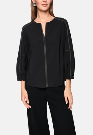 Woman wearing black long-sleeve blouse with white stitching and split neckline, paired with black wide-leg pants on light background.