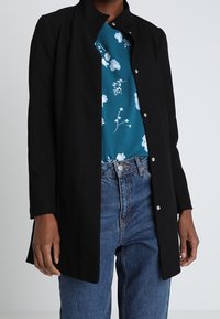 Chaqueta negra de botonadura central con cuello mao, llevada sobre una blusa de flores en color verde azulado y jeans de talle alto en azul. Tejido suave, ajuste entallado.