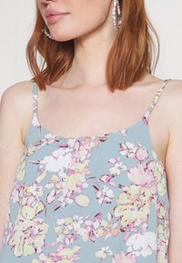 Blomster camisole i lyseblåt stof med justerbare spaghetti-stropper. Har mønstre af pink og gule blomster, glat tekstur.