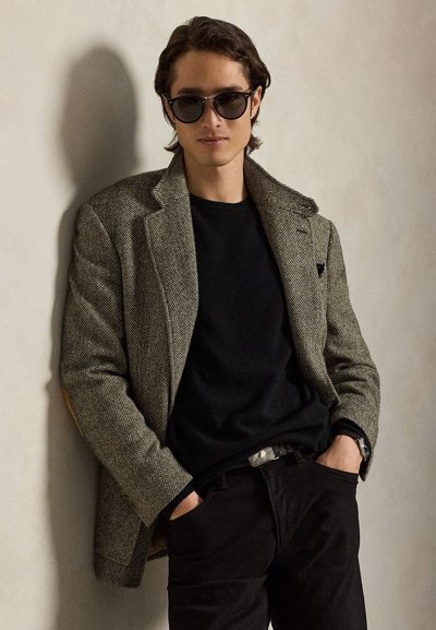 Junger Mann trägt einen gemusterten Blazer, schwarzen Pullover, dunkle Hose und Sonnenbrille, steht mit den Händen in den Taschen vor einer schlichten Wand.