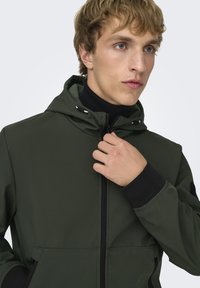 Giacca verde scuro con cappuccio, chiusura frontale con zip, polsini neri e due tasche frontali. Presenta una texture morbida e una vestibilità avvolgente e comoda.