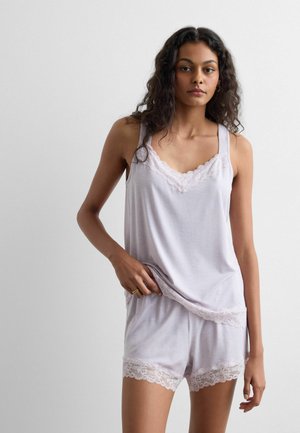 Vrouw met krullend haar, gekleed in een lichtpaarse spaghettitop met kanten randjes en bijpassende korte broek, tegen een effen lichte achtergrond.