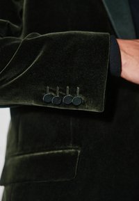 Rękaw ciemnozielonej aksamitnej marynarki z czterema teksturowanymi guzikami, o gładkim wykończeniu i kontrastowym czarnym mankiecie widocznym poniżej.