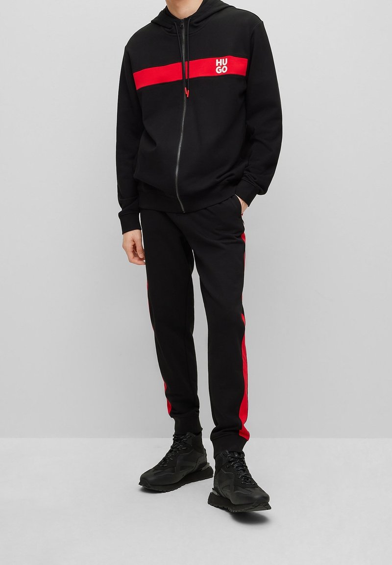 Homme portant un sweat à capuche zippé noir et rouge et un jogging assorti avec des baskets noires, debout devant un fond gris uni.