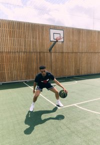 Basketball player in black T-shirt and shorts, dribbling a yellow-black basketball on a green court with wooden walls and a hoop.