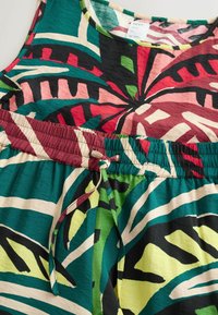 Sukienka bez rękawów o tropikalnym wzorze, z elastycznym pasem i troczkiem, ozdobiona abstrakcyjnymi liśćmi w kolorach zielonym, czerwonym, żółtym, czarnym i kremowym.