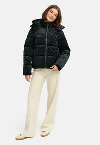 Doudoune noire avec capuche, motif matelassé, fermeture éclair à l'avant et texture douce. Associée à un pantalon de couleur claire et des chaussures décontractées.