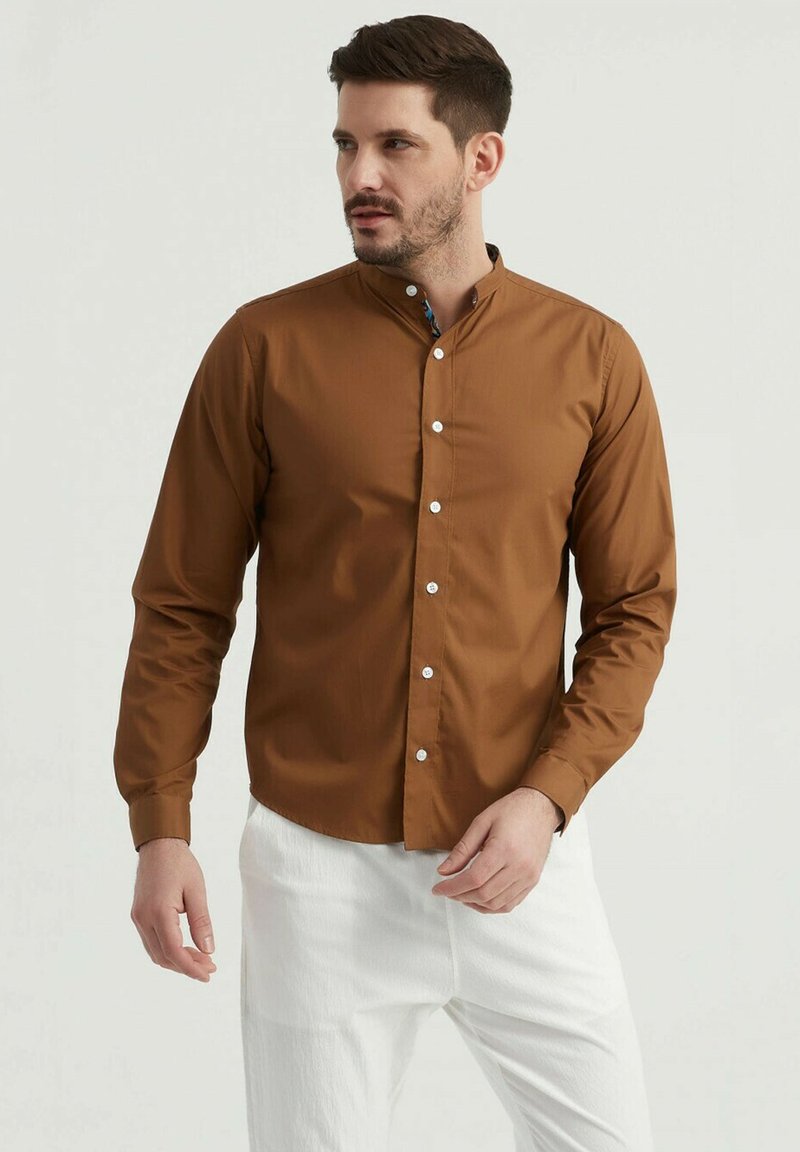 Chemise marron à manches longues avec col italien, dotée de boutons blancs et d'une texture lisse, associée à un pantalon blanc.