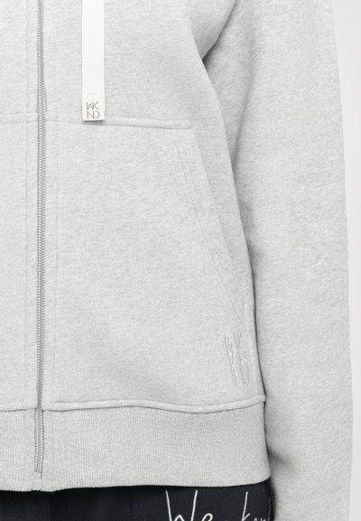 Grå zip-up hettegenser med frontlomme, teksturert stoff og hvit snor. Brodert logodetalj på nederste del.