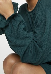 Haut texturé en vert foncé avec des manches volantées et bouffantes. Le tissu semble léger avec un effet froissé. La peau du modèle est brun foncé.