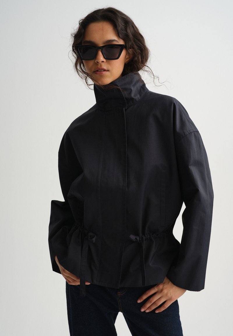 Žena v nadrozmernej čiernej bunde s vysokým golierom a šnúrkou v páse, v tmavých slnečných okuliaroch, stojaca pred jednoduchým pozadím.