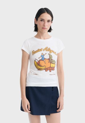 Femme aux cheveux courts et foncés portant un t-shirt blanc avec un motif de fruits et de personnage ainsi qu’un texte, associé à une jupe bleu foncé.
