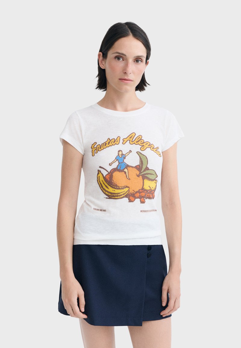 Femme aux cheveux courts et foncés portant un t-shirt blanc avec un motif de fruits et de personnage ainsi qu’un texte, associé à une jupe bleu foncé.