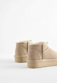 Beige Wildleder-Ankle-Boots mit Futter aus Fell, runder Form und flacher Gummisohle. Auffällige Nähte entlang der Nähte.